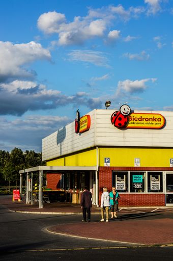 A group of people standing at the front entrance of a Biedronka supermarket with a parking lot