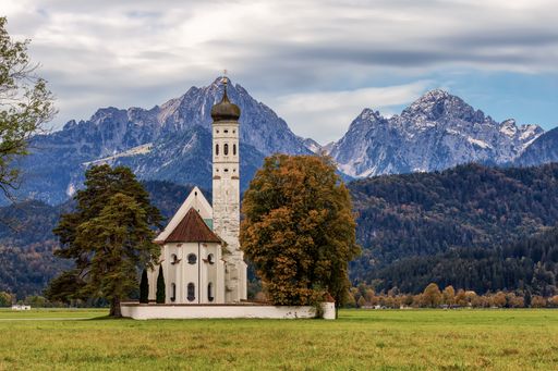 Scenic view of St. Coloman Church with autumn trees and the Bavarian Alps in the background under a cloudy sky.