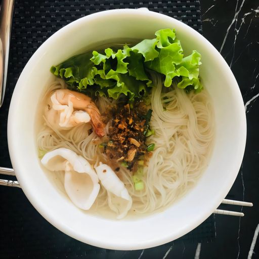 A delicious bowl of Vietnamese noodle soup with shrimp, squid, and fresh greens, served on a black marble table.