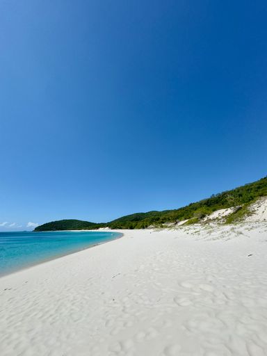 Pristine white sand beach with turquoise waters and lush green hills under a clear blue sky.