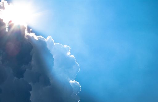 A low angle shot of the sun shining from behind the clouds in a light blue sky