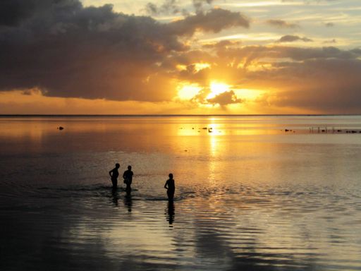 A silhouette of people walking in the sea with the sun shining behind the clouds