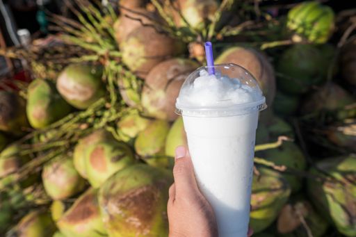 A selective focus shot of a hand holding a coconut juice in a plastic cup with a plastic straw