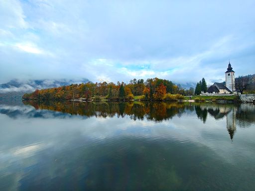Scenic view of Lake Bohinj with autumn trees and a church reflecting in the calm water under a cloudy sky in Slovenia.