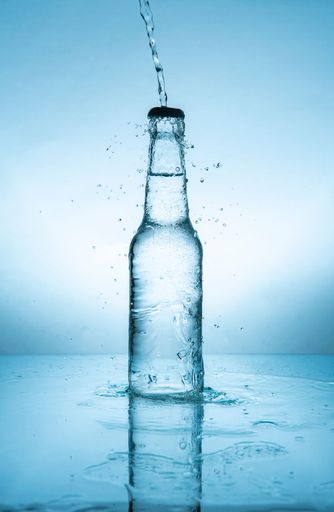Water stream splashing on glass bottle with cap filled with water on wet reflective blue surface against blue background