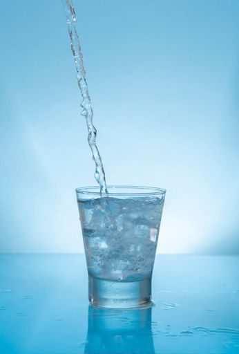 Water stream splashing on glass cup filled with water and ice on wet reflective blue surface against blue background