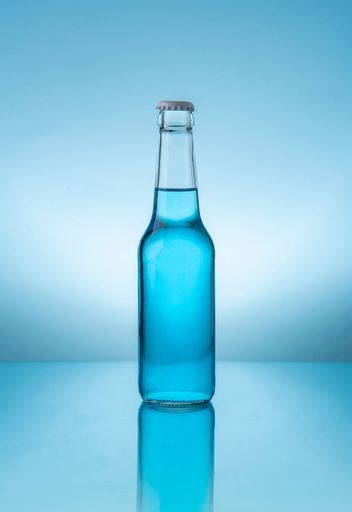 Glass bottle with cap containing blue liquid on reflective blue surface against blue background