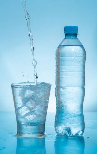 Water stream splashing on frozen glass cup filled with water and ice next to plastic bottle on wet reflective blue surface against blue background