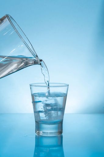 Glass pitcher filling a chilled glass tumbler with ice with water on wet reflective blue surface against blue background