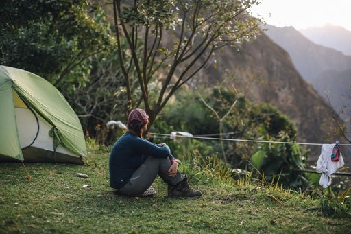 A woman in her 20s sits by a tent, enjoying a serene mountain view at sunset, with lush greenery around.