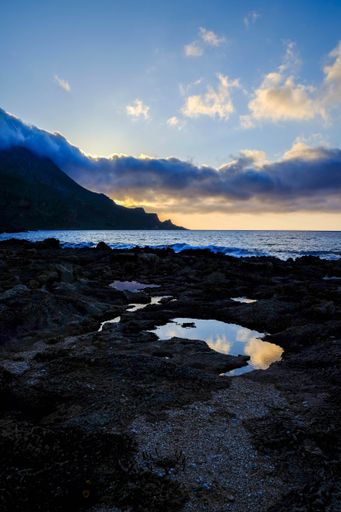 A vertical shot of small ponds in middle off rocks near the sea with mountains in the distance under a blue sky
