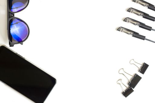 A sunglass, notepad, pens and a smartphone on a white surface viewed from above