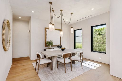 Modern dining room interior with wooden flooring, elegant lighting, and large windows overlooking greenery.