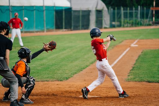 A youth baseball player in orange jersey swings bat, hitting ball into outfield