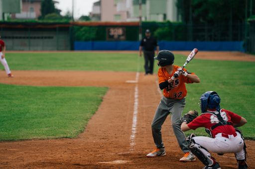 A youth baseball player in orange jersey swings bat, hitting ball into outfield