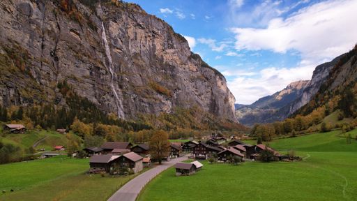 Scenic view of Lauterbrunnen Valley with waterfall, lush green fields, and traditional Swiss chalets under a partly cloudy sky.