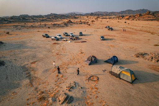 Aerial view of a desert camping site with tents and vehicles, surrounded by rocky terrain under a clear sky.
