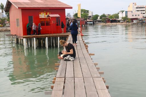 Profile view from a sitting Caucasian young woman, sitting on a narrow wooden pier and using her smartphone at Georgetown harbor in Malaysia.
