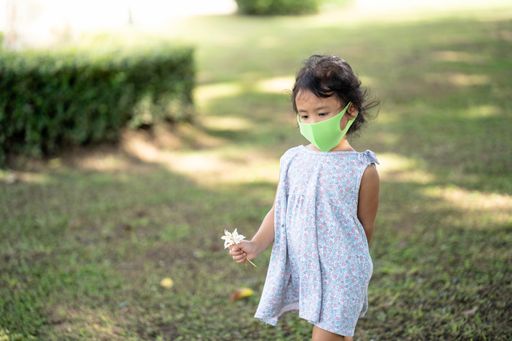 A cute little Thai girl in a protective face mask carrying a white flower walking in a park