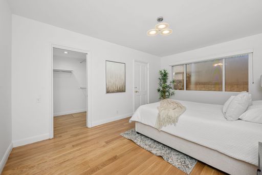 Modern minimalist bedroom with wooden flooring, white walls, cozy bed, and large window, featuring a walk-in closet.