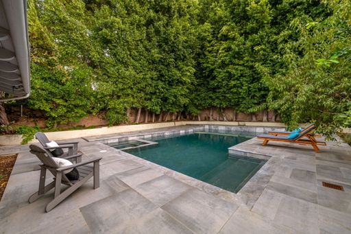 A modern backyard with a swimming pool, lounge chairs, and lush green trees