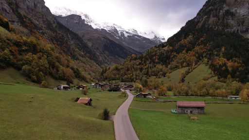 Scenic view of a rural mountain village nestled in a lush green valley with snow-capped peaks under a cloudy sky.