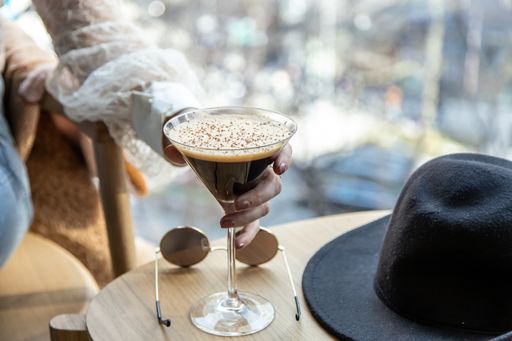 close up view of a woman's hand picking a martini glass filled with delicious chocolate coffee martini. sunglasses and black hat on coffee table
