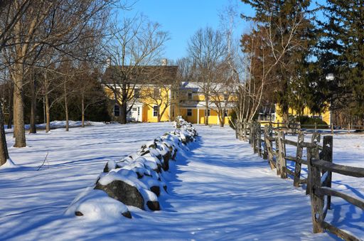 A closeup shot of a yellow house surrounded by trees and a road covered in snow in Hamilton MA