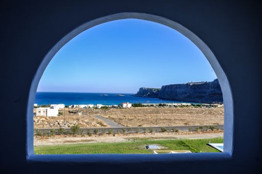 An arched type window with a view of the road in the middle of dry grass fields near the sea with blue sky in the background