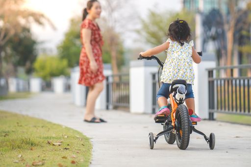 A back view of a cute little Thai girl riding a bicycle in a park with her mother in the background