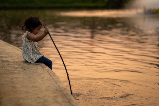 A little Thai girl sitting holding a wooden stick playing near the pond at sunset