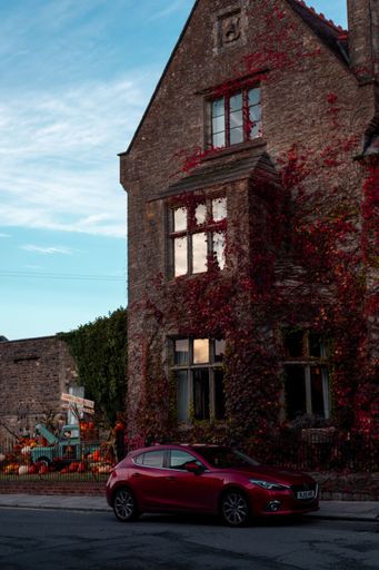A red car parked in front of a historic stone building covered with red ivy, under a clear blue sky in a quaint village setting.