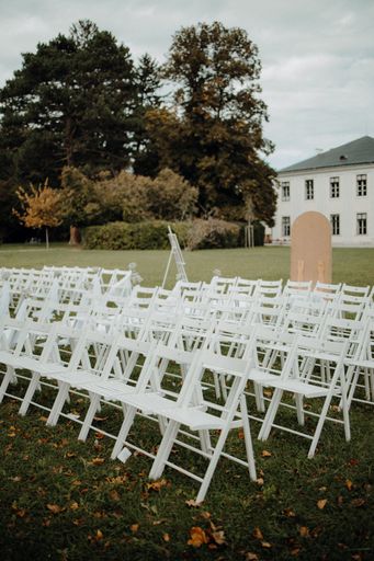 Outdoor wedding setup with rows of white chairs on a grassy lawn, surrounded by lush trees and a historic building in the background.