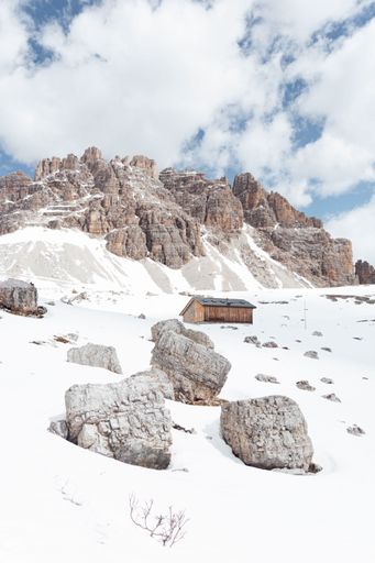 A small wooden cabin rests in a snow-covered landscape with rocky mountains in the background.