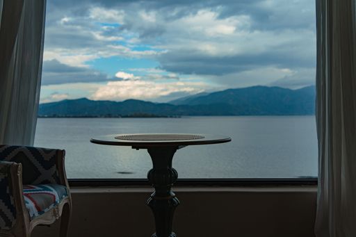 Scenic view of a lake and mountains through a window with a table and chair in the foreground, under a cloudy sky.