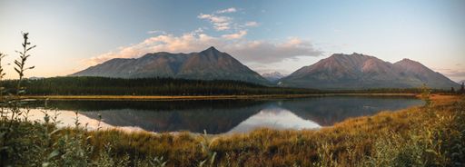Panoramic view of serene lake reflecting mountains under a blue sky at sunset, surrounded by lush greenery.