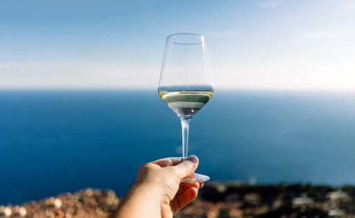 Hand holding glass of white wine with beach on background - Travel vacation concept