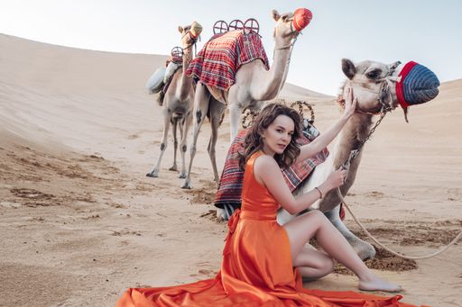 A gorgeous Caucasian woman wearing an orange long dress sitting on the sand with camels in a desert