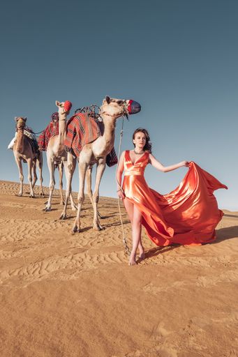 A gorgeous Caucasian woman wearing an orange long dress standing in front of camel in a desert