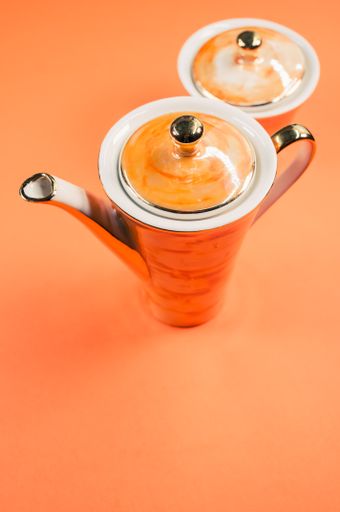 A close up of an orange covered teapot, a covered cup on an orange isolated background with free space