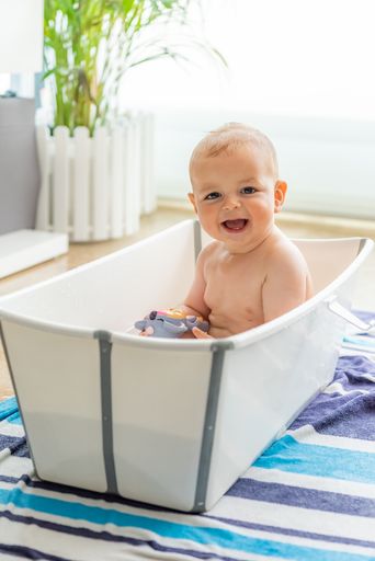 A caucasian baby chewing on a toy and looking out of the corner of his eye that is tucked inside a portable bathtub in the living room