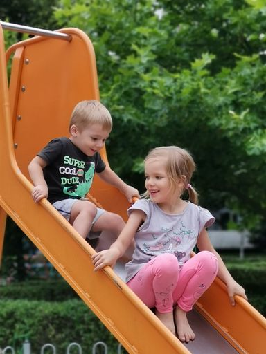 A vertical shot of two blond children, male and female, playing on an orange slide, Pol