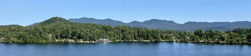 Panoramic view of a serene lake surrounded by lush green forests and distant mountains under a clear blue sky.