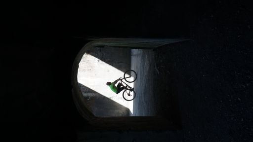 A vertical distant shot through a stone arch of a cyclist under a beam of sunlight