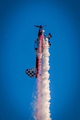 An aerobatic airplane performing a vertical climb with smoke trail against a clear blue sky.
