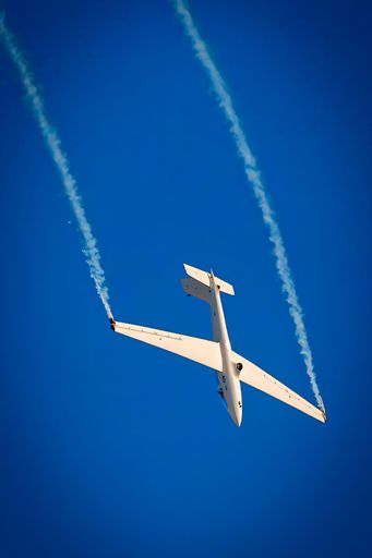 A glider performs an aerobatic maneuver against a clear blue sky, leaving smoke trails in its wake.