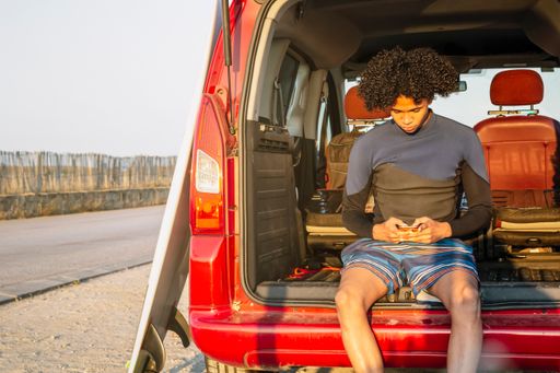 A young, latino surfer with afro hair chatting on a mobile phone sitting  in the car trunk at sunset