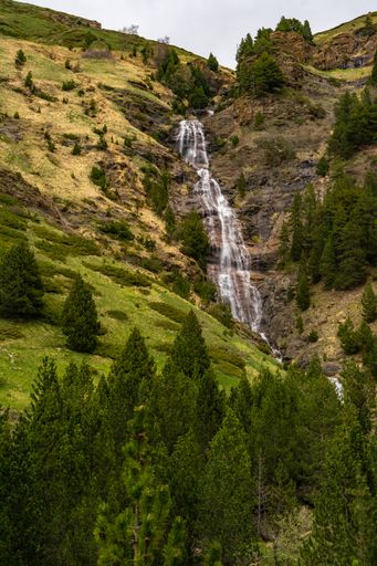 A vertical shot of a landscape with a waterfall from a mountain with green trees and fields