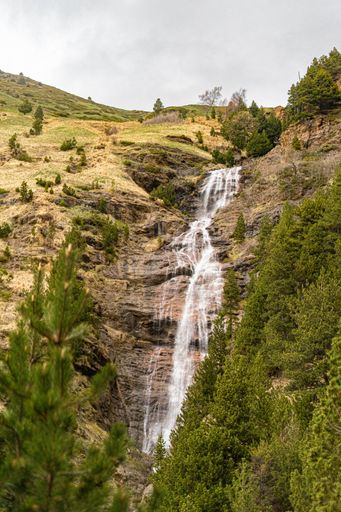 A vertical shot of a landscape with a waterfall from a mountain with green trees and fields