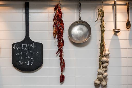 A pan, wooden peel, metal soup spoon, dried red pepper, and garlic hanging from a white wall